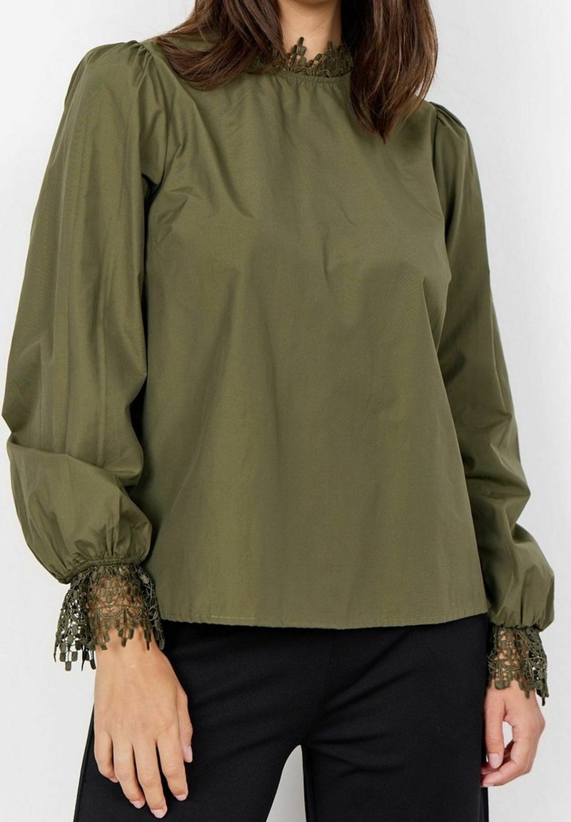 Femme portant un chemisier vert olive à manches longues avec une bordure en dentelle au col et aux poignets, associée à un pantalon noir, debout devant un fond blanc.