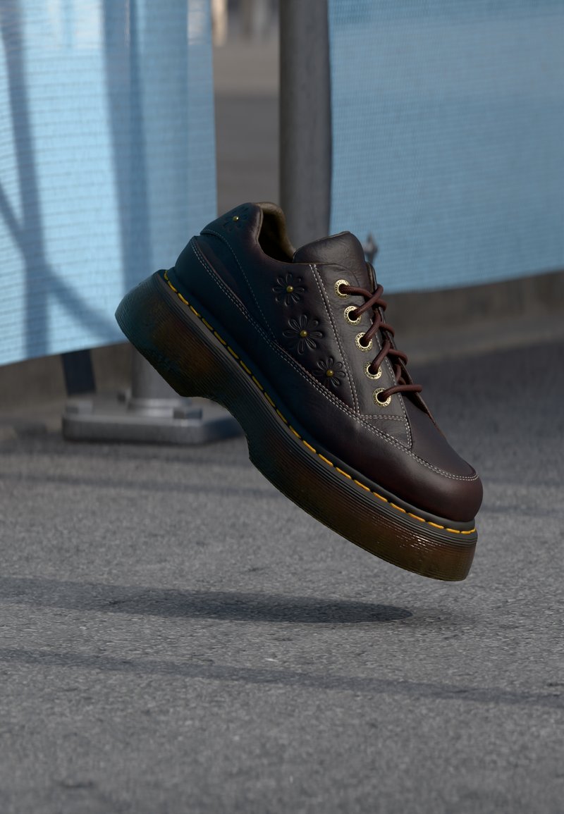 Zapato plataforma de cuero marrón oscuro con estampados florales, costuras amarillas y suela gruesa translúcida flotando sobre una calle de asfalto.