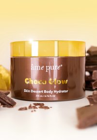 Brun burk med ett guldlock märkt "Choco Glow", innehållande kroppshydrator, med en glänsande yta och omgiven av chokladbitar.