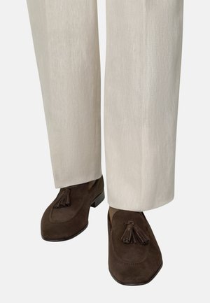 Brązowe mokasyny zamszowe z frędzlami, noszone z jasnobeżowymi spodniami o prostych nogawkach na jednolitym tle.