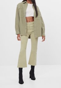 Camisa oversized verde oliva sobre una blusa corta blanca, combinada con jeans acampanados verde claro y botines negros de tacón. Detalles de tejido texturizado visibles.