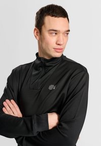 Schwarzes, langärmliges Sportoberteil mit halbem Reißverschluss, strukturiertem Stoff und kleinem, gemustertem Logo auf der Brust.