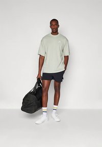 Heleroheline suurem T-särk, mustad lühikesed püksid, valged tossud ja mitmevärvilised triibulised sokid. Modell hoiab suurt musta spordikotti.