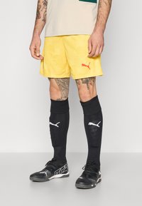 Gule idrettsshorts med et strukturert mønster og en rød Puma-logo. Svarte knelange sokker med hvite Puma-detaljer, kombinert med svarte joggesko.