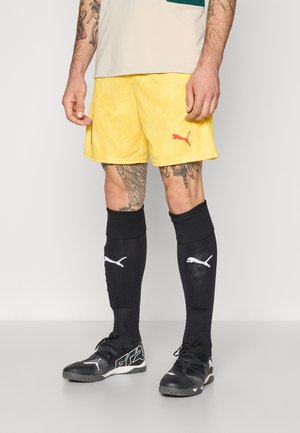 Gule atletiske shorts med et struktureret mønster og et rødt Puma-logo. Sorte knæhøje sokker med hvide Puma-detaljer, kombineret med sorte sportsko.