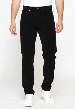 Uomo con pantaloni di velluto a coste neri e sneakers bianche, in piedi davanti a uno sfondo bianco semplice.