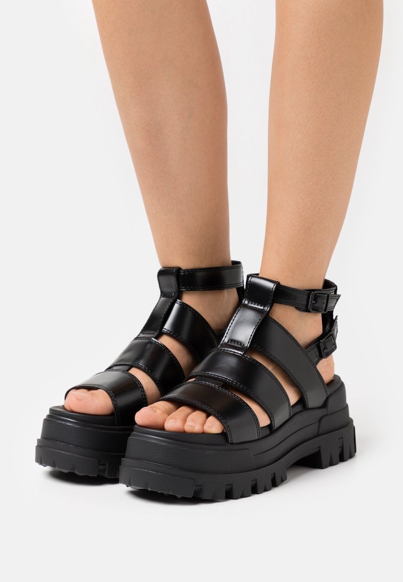 Pieds portant des sandales à plateforme épaisses noires avec plusieurs brides et boucles sur un fond blanc uni.