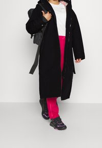 Svart lång kappa med dragkedja, bärs över en vit skjorta och ljusrosa byxor. Grå ryggsäck och svarta skor med rosa detaljer.