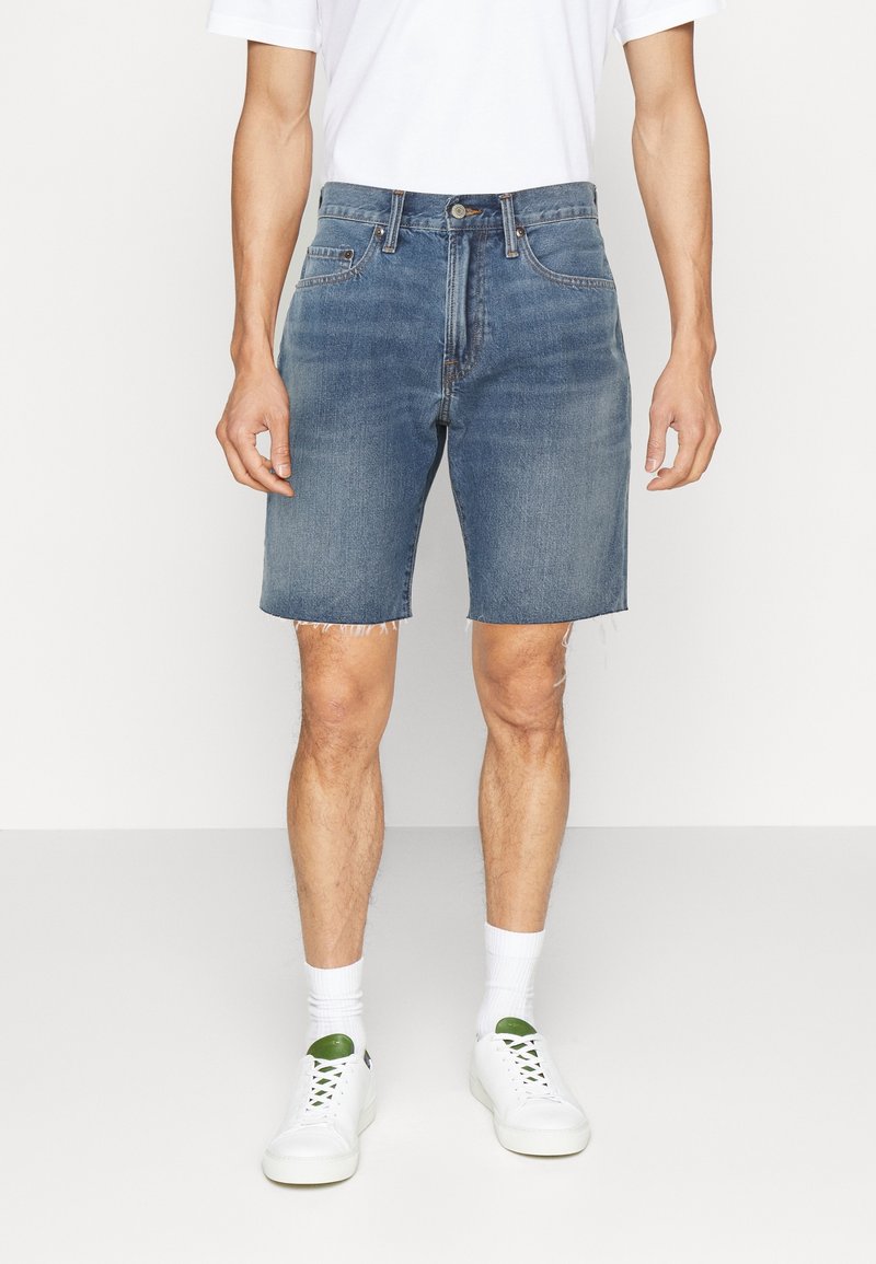 Shorts en jean bleu clair, avec une coupe droite et un ourlet effiloché. Portés avec un t-shirt blanc et des baskets blanches à accents verts.