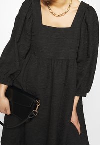 Svart strukturklänning med fyrkantig halsringning och puffärmar, tillsammans med en svart kuvertväska med guldiga metalldetaljer.