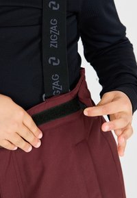 Pantaloni bordeaux con bretelle nere a zigzag regolabili, dotati di chiusura in velcro in vita e una texture di tessuto liscia.