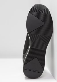 Semelle de sneaker noire avec rainures texturées et motifs triangulaires, affichant la marque Michael Kors au niveau du talon.