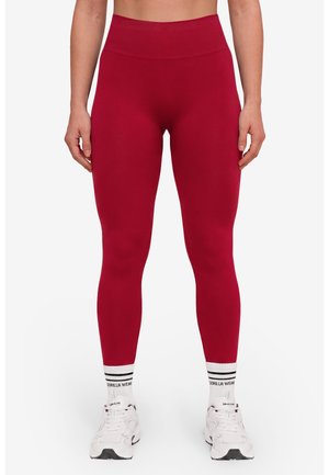 Persoon draagt hoge rode leggings met witte sportsokken en witte sneakers, staand tegen een effen achtergrond.
