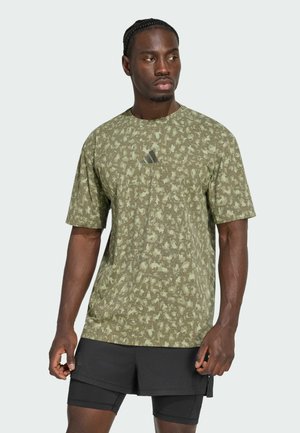 Mand i grøn atletisk tshirt med mønster og sorte lagdelte shorts står med afslappede hænder og kigger let til siden mod en ensfarvet baggrund.