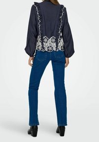 Femme debout, de dos, portant un chemisier bleu marine avec des volants et de la dentelle blanche à motifs floraux, un jean bleu et des chaussures noires à talons.