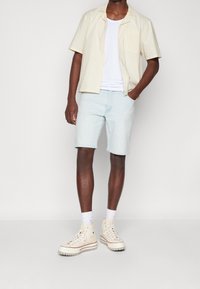 Světle barevná košile s krátkým rukávem, bílý tílko, světle modré šortky a béžové vysoké tenisky s červenými akcenty a bílými ponožkami.