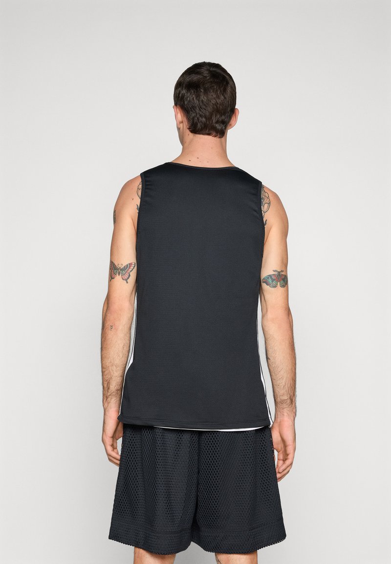 Canottiera sportiva nera con una finitura testurizzata, caratterizzata da accenti laterali bianchi. Indossata con pantaloni corti in rete neri, che mettono in mostra tatuaggi sulle braccia.