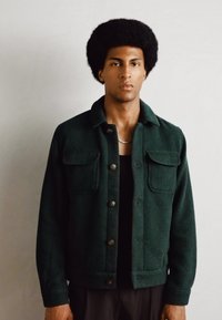 Giacca di velluto a coste verde con due tasche sul petto, chiusura con bottoni e texture a coste, indossata sopra una maglietta nera.