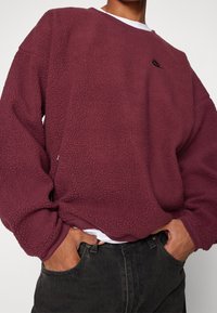 Sudadera de forro polar burdeos con acabado texturizado, cuello redondo y detalle de logo negro. Presenta mangas con puños y un ajuste holgado.