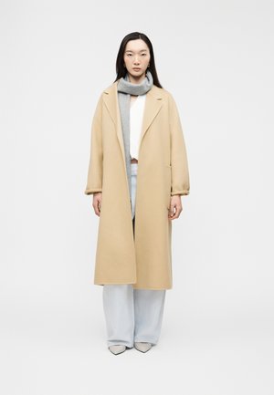 Γυναίκα που φοράει μακρύ μπεζ παλτό, γκρι κασκόλ, λευκό τοπ, ανοιχτόχρωμο φαρδύ παντελόνι και μυτερές λευκές γόβες, στέκεται μπροστά σε απλή φόντο.