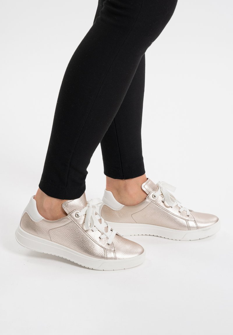 Metallic roségoldene Sneakers mit weißen Akzenten, strukturiertem Obermaterial und weißen Schnürsenkeln, kombiniert mit schwarzen eng anliegenden Hosen, vor einem weißen Hintergrund.