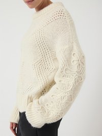 Frau, die einen cremfarbenen, grob gestrickten Pullover mit strukturierten Zickzackmustern und floralen Kabelstrickdetails an den Ärmeln trägt.