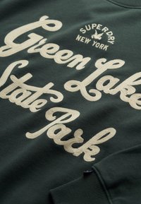 Sudadera de color verde oscuro con texto bordado color crema que dice "Green Lake State Park" y "Superdry New York" en fuente cursiva.
