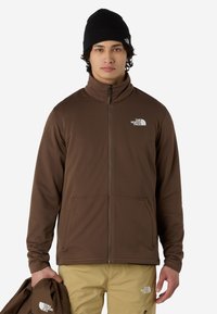 Brązowa kurtka na zamek z teksturowanej tkaniny, z wysokim kołnierzem, dwoma przednimi kieszeniami i białym logo na lewej piersi.