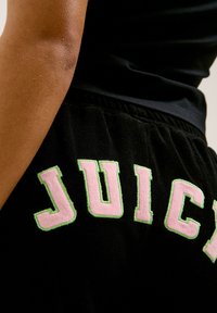 Jogging noir en tissu doux avec une surface texturée, arborant "JUICY" en lettres brodées roses et vertes à l'arrière.