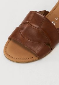 Sandal in pelle marrone a scivolo con design a fascia curva. La suola è di colore beige con dettagli di cucitura, presenta un'apertura per le dita e un profilo minimalista.