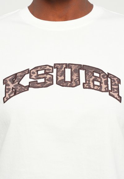 Balta kokvilnas t-krekls ar "KSUBI" logotipu brūnā kamuflāžas motīvā, ar apaļu apkakli un īsām piedurknēm. Dekoratīvas šuvju akcenti.