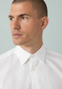 B�ílá košile s knoflíky a strukturovaným límcem, vyrobená z hladké a lehké tkaniny s klasickým střihem. Detailní pohled zdůrazňuje texturu.
