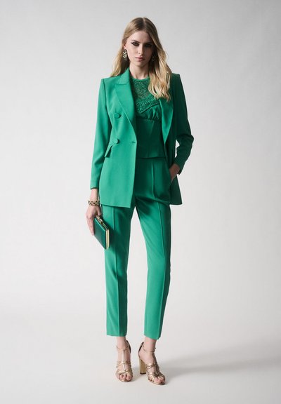 Completo su misura verde con giacca doppiopetto e pantaloni attillati. Top verde con texture sotto. Abbinato a sandali con tacco dorati e una pochette.