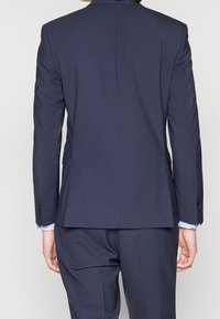 Mörkblå kostymjacka med skräddarsydd design, enkel slits bak och matchande byxor. Blå skjortmanschetter syns vid handlederna.