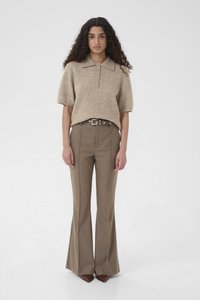 Frau mit langen lockigen Haaren, die ein beiges, kurzärmeliges Strick-Poloshirt, braune ausgestellte Hosen mit Gürtel und spitze braune Schuhe trägt, steht vor weißem Hintergrund.