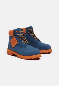 Stivali in suede blu con un collare imbottito arancione, lacci arancioni e una suola robusta arancione. Presentano un'etichetta con il marchio sul lato.