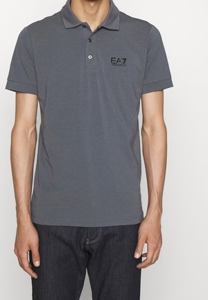Hombre con una camisa polo de manga corta color gris oscuro con el logotipo de EA7 Emporio Armani en el pecho izquierdo y jeans oscuros.