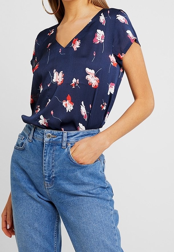 Femme portant un chemisier bleu marine à col en V avec un imprimé floral rose et blanc, rentré dans un jean taille haute bleu clair, main dans la poche.