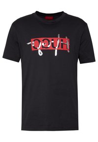 Black cotton t-shirt featuring a bold, slanted red rectangle with the word "HUGO" in silver metallic finish and white accents. Short sleeves, crew neck.
