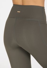 Hooggetailleerde, donkergroene legging met een soepele textuur, voorzien van contrasterende stiksels en een klein metalen logo op de achterkant.