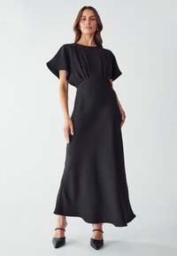 Robe maxi noire avec un corsage ample et une taille froncée. Texture douce, manches courtes évasées et jupe fluide avec un léger éclat.