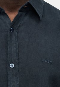 Κοντινό πλάνο σε γιακά σκούρου μπλε πουκάμισου με κουμπιά και κεντημένο «BOSS» στο αριστερό στήθος, που φοριέται από άτομο με σκούρο δέρμα.