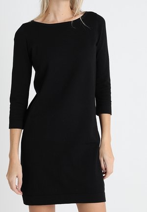 Femme portant une robe noire simple, longueur genoux, avec des manches trois-quarts, sur un fond clair uni.