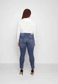 Blouse blanche à manches longues avec un col à volants, associée à un jean skinny bleu marine avec des poches arrière visibles et des escarpins noirs.