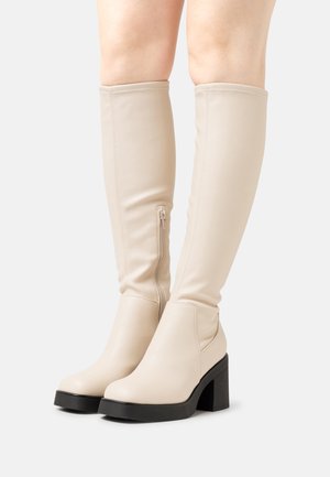 Botas de imitação de couro bejes até ao joelho com um salto preto robusto, apresentando um fecho lateral e uma textura lisa e suave.