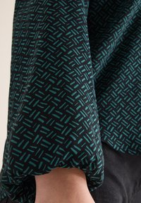 Schwarze Stoffbluse mit türkisfarbenem geometrischen Muster. Sie hat einen lockeren Schnitt und versammelte Ärmeldetails, die die Textur und die Schlichtheit des Designs betonen.