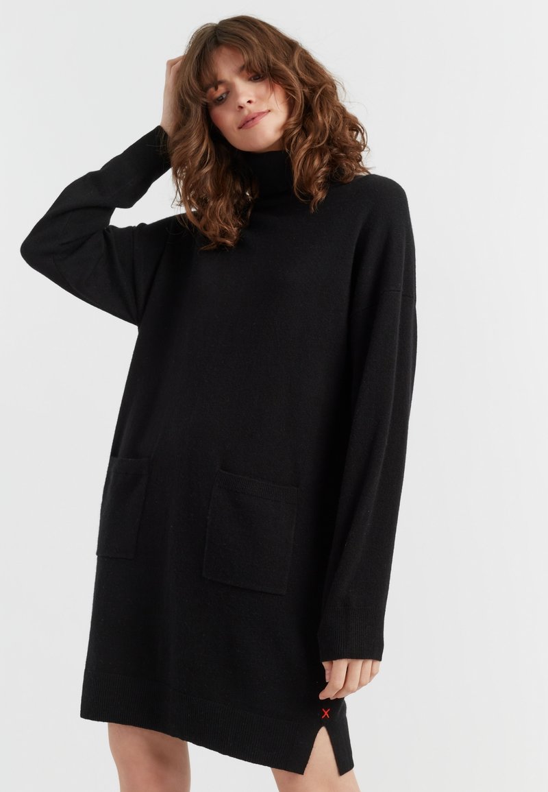 Schwarzes Strickkleid mit hohem Kragen, langen Ärmeln, zwei Fronttaschen, seitlichen Schlitzen und roten Akzentstichen am Saum.