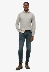 Grau gestrickter Pullover über einem hellen Kragenhemd, blaue Jeans und braune Schnürstiefel. Das Modell steht frontal.