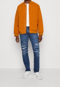 Veste bombardier orange, t-shirt blanc, jeans bleus déchirés et baskets blanches. La veste possède une fermeture éclair et des poignets côtelés.