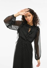 Robe noire à pois blancs avec des manches bouffantes transparentes et un col à volants. Le tissu est léger avec une silhouette fluide et une fermeture à nouer au col.